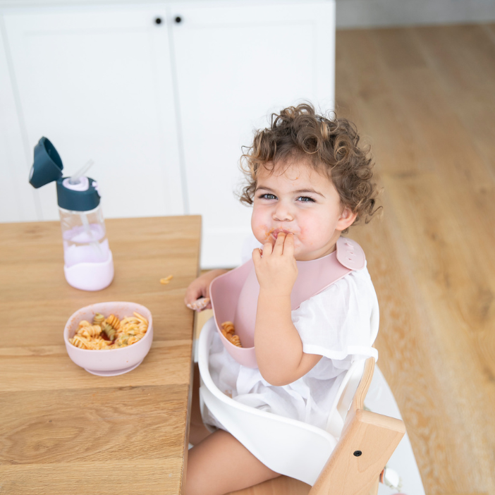 Toddler eating pasta with pink mess-free bib at wooden table, food-safe catch-all design