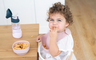 Toddler eating pasta with pink mess-free bib at wooden table, food-safe catch-all design