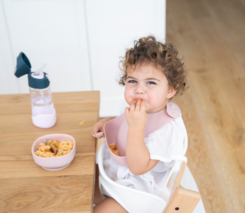 Toddler eating pasta with pink mess-free bib at wooden table, food-safe catch-all design