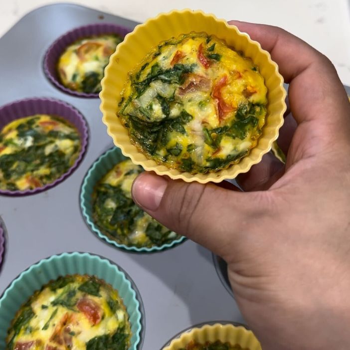 Hand holding yellow silicone egg muffin cup with spinach and tomato, baked in muffin tin