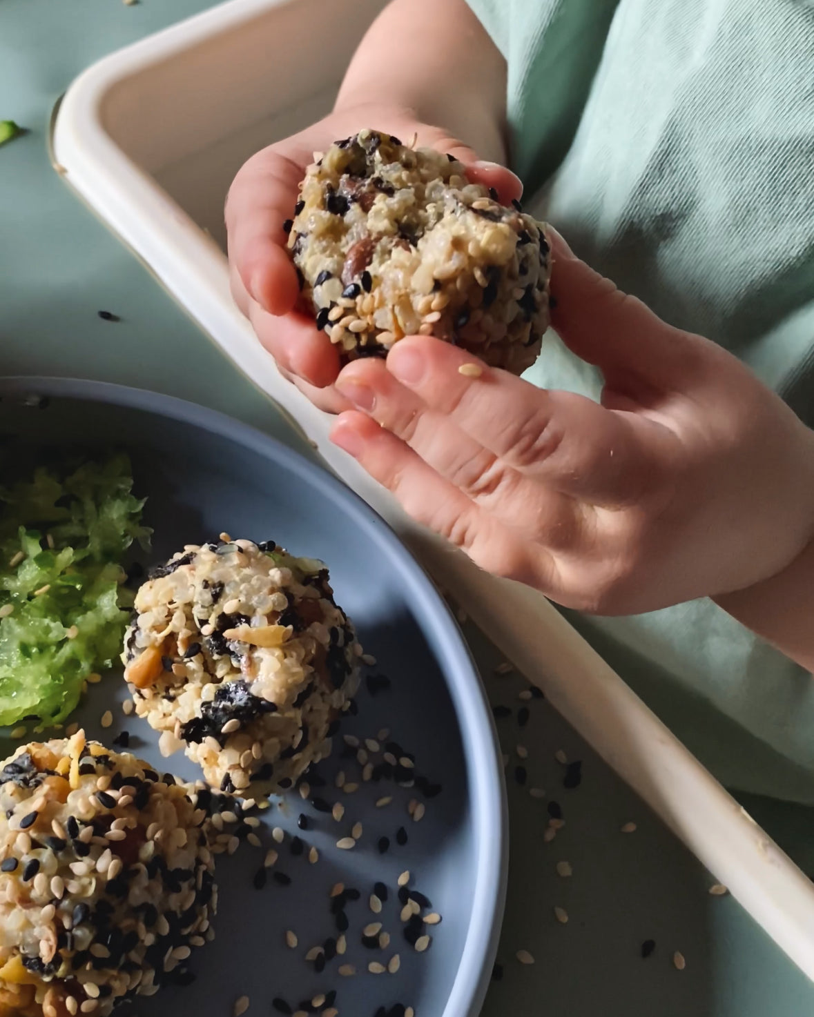 Child's hands holding sesame-coated rice ball over plate, mealtime mess-free with catch tray