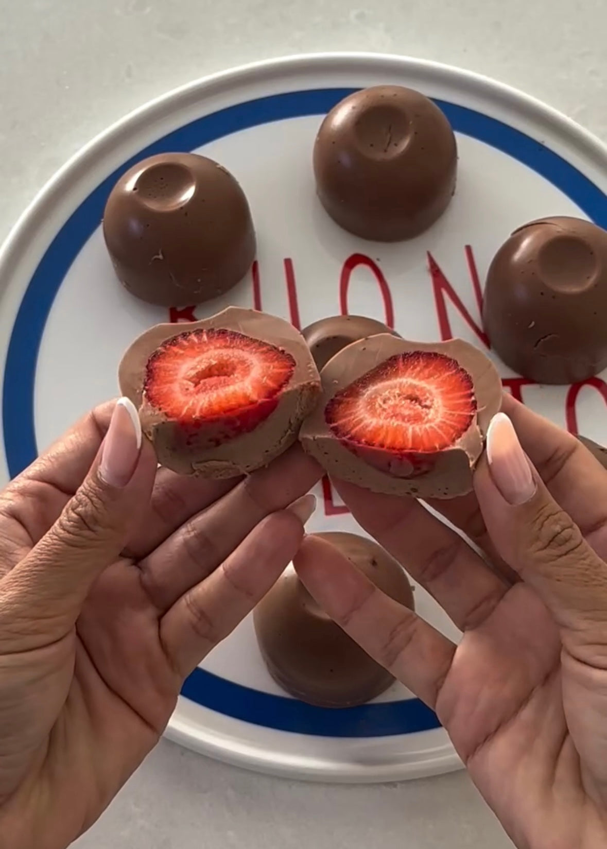 Hands holding chocolate-covered strawberry halves, revealing fresh red interior, with whole chocolates on plate