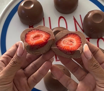 Hands holding chocolate-covered strawberry halves, revealing fresh red interior, with whole chocolates on plate