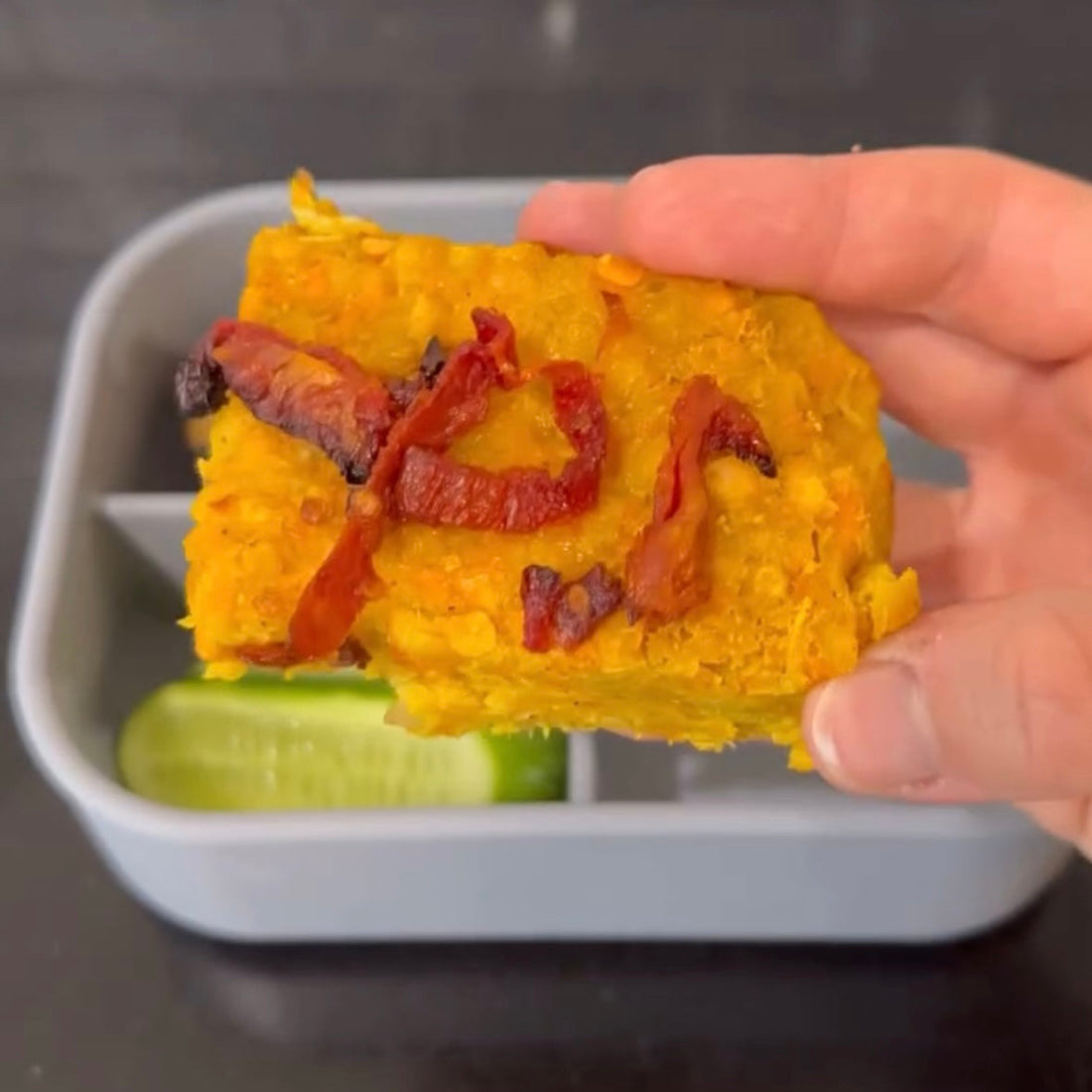 Hand holding savory yellow baked dish with sun-dried tomatoes over bento box with cucumber slices