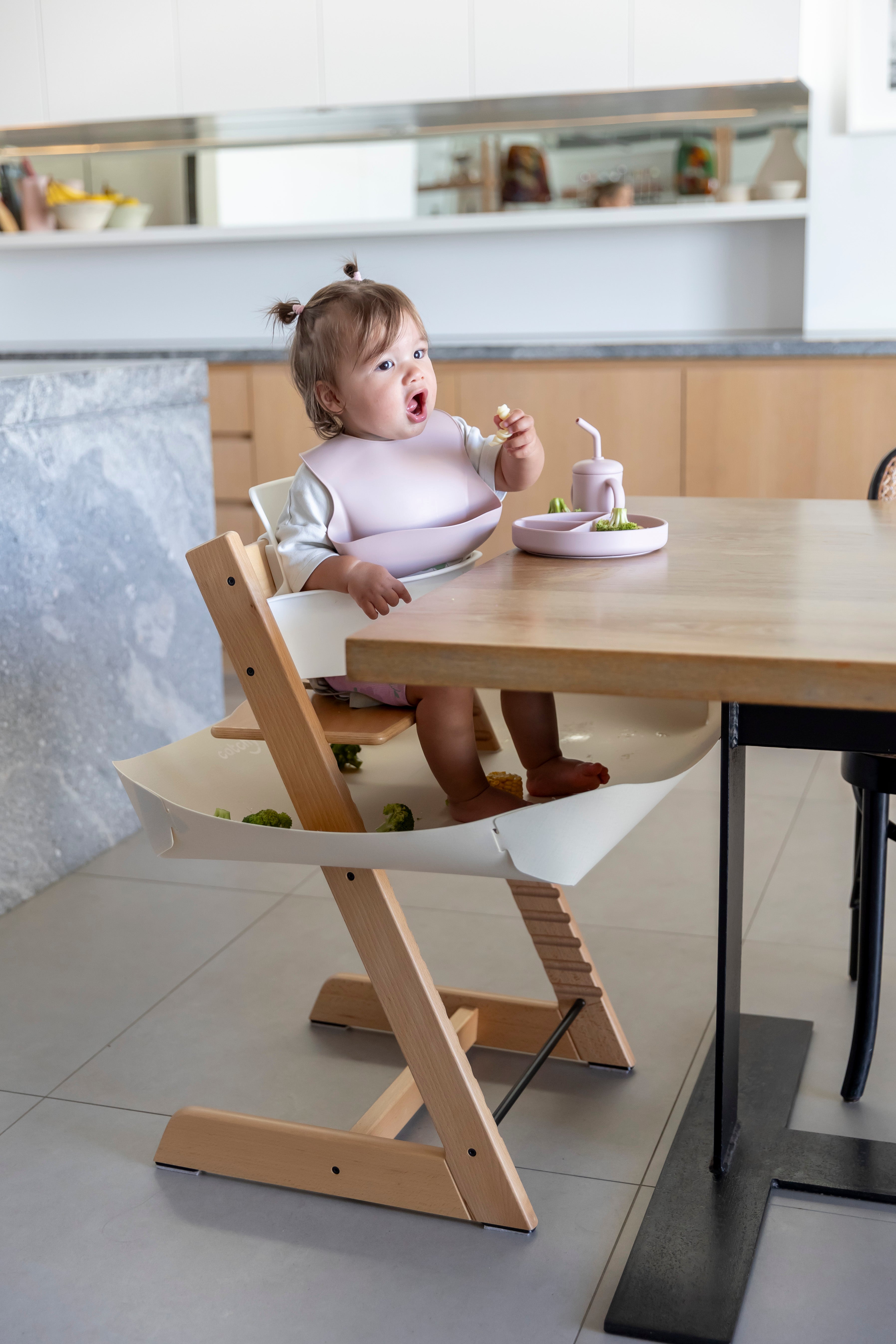 Baby eating with mess-free bib and food catcher under high chair, protecting floors during mealtime.
