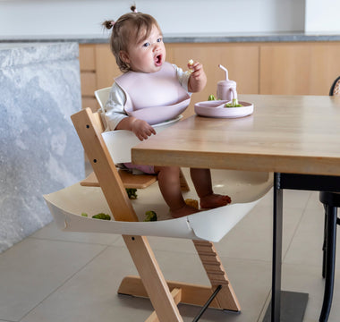 Baby eating with mess-free bib and food catcher under high chair, protecting floors during mealtime.